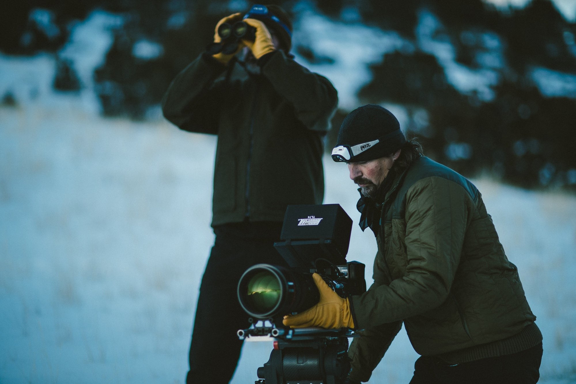 A wildlife film crew working in the snow with a cinema camera and binoculars